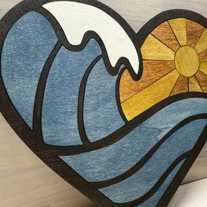 May include: Wooden heart-shaped wall art featuring a wave and sun design. The wave is blue and white, the sun is yellow and orange, and the outline is dark brown. A decorative piece for home decor.