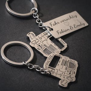 Puede incluir: Llaveros plateados con diseño de camión. Un llavero tiene una etiqueta rectangular con el texto "Fahr vorsichtig" y "Fabian & Emily" grabado. Los llaveros están conectados a un anillo plateado.