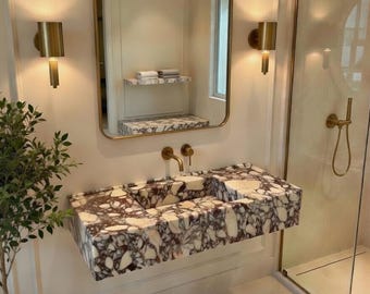 Pode incluir: Uma casa de banho moderna com uma bancada, um espelho retangular e acessórios dourados. A bancada é feita de pedra com um padrão branco e roxo. Duas arandelas douradas ladeiam o espelho. Uma pequena planta em vaso é visível.