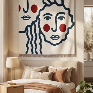 K&ouml;nnte beinhalten: Eine gro&szlig;e, cremefarbene Wandtapisserie mit einem minimalistischen Design von zwei Gesichtern mit marineblauen Umrissen und roten Akzenten. Sie h&auml;ngt &uuml;ber einem Bett mit neutralfarbener Bettw&auml;sche und Kissen. Ein Fransenrand sorgt f&uuml;r Textur.