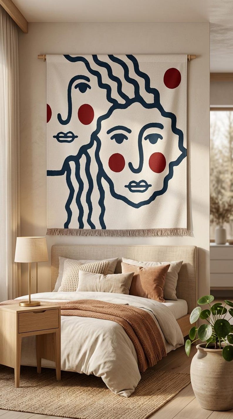K&ouml;nnte beinhalten: Eine gro&szlig;e, cremefarbene Wandtapisserie mit einem minimalistischen Design von zwei Gesichtern mit marineblauen Umrissen und roten Akzenten. Sie h&auml;ngt &uuml;ber einem Bett mit neutralfarbener Bettw&auml;sche und Kissen. Ein Fransenrand sorgt f&uuml;r Textur.