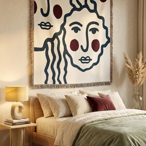 K&ouml;nnte beinhalten: Eine cremefarbene Wandtapisserie mit einem minimalistischen Gesichtsdesign in Marineblau und Burgunderrot. Das Kunstwerk h&auml;ngt &uuml;ber einem Bett mit cremefarbener Bettw&auml;sche und einer salbeigr&uuml;nen Decke. Ein Beistelltisch mit einer Lampe steht neben dem Bett.