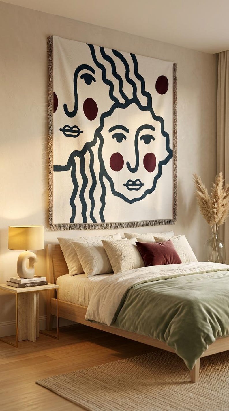 K&ouml;nnte beinhalten: Eine cremefarbene Wandtapisserie mit einem minimalistischen Gesichtsdesign in Marineblau und Burgunderrot. Das Kunstwerk h&auml;ngt &uuml;ber einem Bett mit cremefarbener Bettw&auml;sche und einer salbeigr&uuml;nen Decke. Ein Beistelltisch mit einer Lampe steht neben dem Bett.