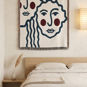 K&ouml;nnte beinhalten: Eine cremefarbene Wandtapisserie mit zwei stilisierten Gesichtern mit marineblauen Umrissen und burgunderfarbenen Akzenten. Die Tapisserie ist rechteckig mit einem Fransenrand und h&auml;ngt &uuml;ber einem Bett mit beiger Bettw&auml;sche.