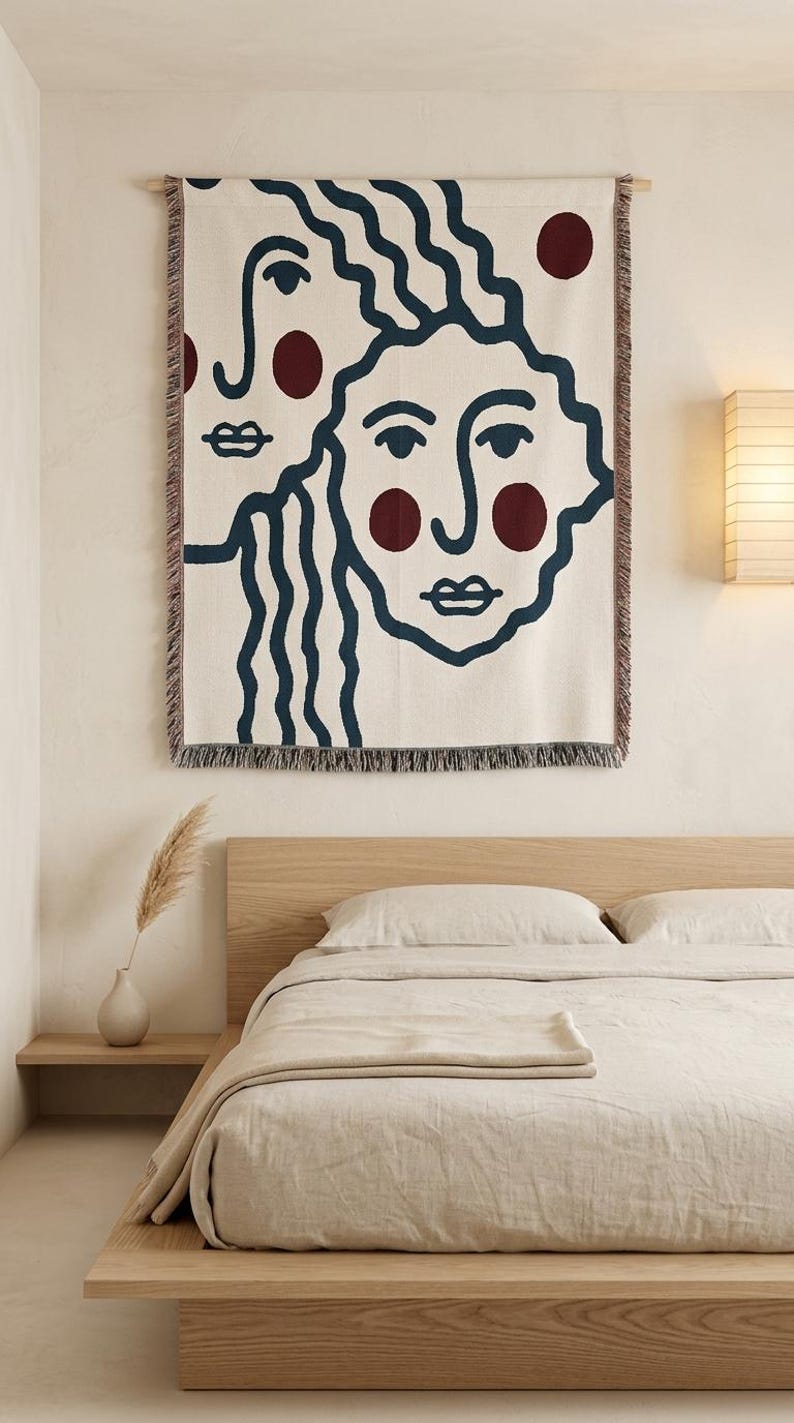 K&ouml;nnte beinhalten: Eine cremefarbene Wandtapisserie mit zwei stilisierten Gesichtern mit marineblauen Umrissen und burgunderfarbenen Akzenten. Die Tapisserie ist rechteckig mit einem Fransenrand und h&auml;ngt &uuml;ber einem Bett mit beiger Bettw&auml;sche.