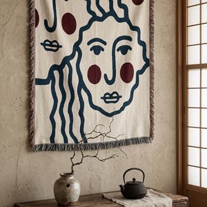 K&ouml;nnte beinhalten: Eine cremefarbene gewebte Wurfadecke mit einem minimalistischen Gesichtsdesign in Marineblau und Burgunderrot. Die Decke zeigt zwei stilisierte Gesichter mit welligem Haar und kreisf&ouml;rmigen Akzenten. Die Kanten haben eine Fransenborte.