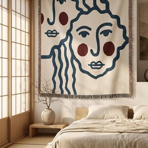 K&ouml;nnte beinhalten: Ein gro&szlig;er, cremefarbener Wandteppich mit zwei abstrakten Gesichtern mit blauen Umrissen und weinroten Akzenten. Der Wandteppich hat einen Fransenrand und h&auml;ngt &uuml;ber einem Bett in einem minimalistischen Schlafzimmer. Der Raum hat Holzakzente und eine neutrale Farbpalette.
