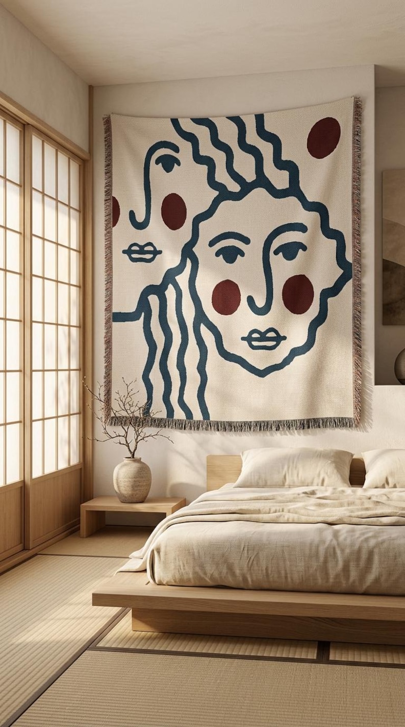 K&ouml;nnte beinhalten: Ein gro&szlig;er, cremefarbener Wandteppich mit zwei abstrakten Gesichtern mit blauen Umrissen und weinroten Akzenten. Der Wandteppich hat einen Fransenrand und h&auml;ngt &uuml;ber einem Bett in einem minimalistischen Schlafzimmer. Der Raum hat Holzakzente und eine neutrale Farbpalette.
