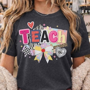 Op de afbeelding: Donkergrijs t-shirt met een kleurrijke afbeelding met de tekst "TEACH". Het ontwerp bevat een hart, een strik met een appel, een discobalhart en stervormen. Het woord "TEACH" is in een mix van lettertypen en kleuren.