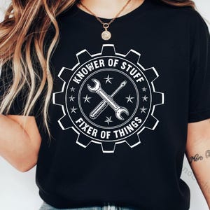 Puede incluir: Camiseta negra con un gráfico blanco. El gráfico es una forma de engranaje con el texto "KNOWer OF STUFF" y "FIXER OF THINGS". Dentro del engranaje hay llaves y un destornillador cruzados, con estrellas.