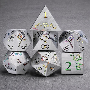 May include: A set of six silver-colored polyhedral dice for tabletop games. Each die features etched numbers and floral designs with colorful accents. The dice are arranged in a balanced composition against a gray background.
