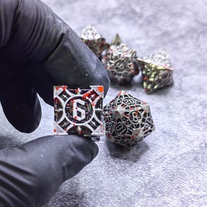 May include: A set of metallic polyhedral dice featuring a silver-toned finish with intricate cut-out details and red splatter accents. The dice display black numbers, including a prominent six on the square die and a twenty on the icosahedron.