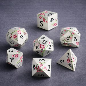 Puede incluir: Un juego de siete dados poliédricos en blanco con números negros y un diseño de ramas de cerezo en flor pintado a mano en rosa. El set incluye un d20, d12, d10, d8, d6, d4 y un dado de porcentaje, todos con motivos florales y números negros.