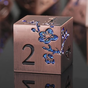 May include: A six-sided metal die with a brushed copper finish. The face displays the number two in a black sans-serif font. A floral branch design is engraved and filled with iridescent blue glitter, extending across the top and side edges.