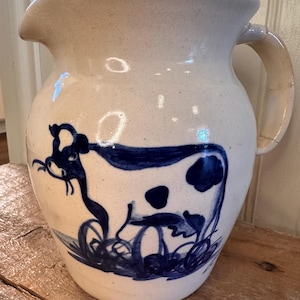 May include: Cream-colored ceramic pitcher with a blue cow illustration. The pitcher has a spout and a curved handle. The cow design features dark blue spots and is set against a white background. The pitcher is sitting on a wooden surface.
