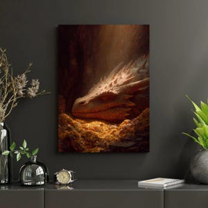 Op de afbeelding: Een canvas print met een slapende draak die rust op een stapel gouden munten. De draak heeft een roodbruine huidskleur met scherpe, stekelige schubben. Het kunstwerk speelt zich af in een donkere grot met lichtstralen.