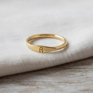 Puede incluir: Un anillo dorado con una cara ovalada y plana. El anillo está grabado con la letra "A". El anillo está sobre un fondo de tela blanca.