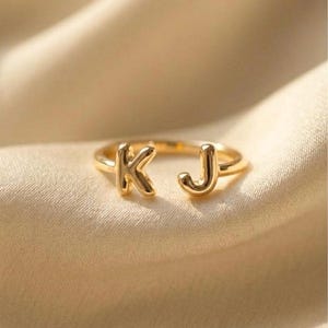 Pode incluir: Um anel dourado com as letras "K" e "J". O anel está colocado sobre um tecido macio de tom neutro. As letras estão em uma fonte simples e elegante, e o anel parece ser feito de metal polido.