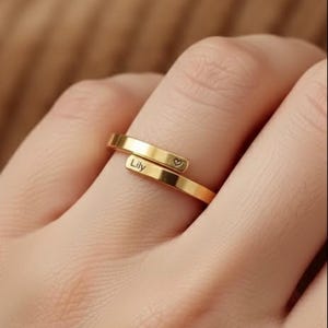 Peut inclure: Une bague dorée avec un design enveloppant. La bague porte le nom "Lily" gravé d'un côté et un petit symbole de cœur de l'autre. La bague est portée sur un doigt.