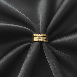 Peut inclure: Une bague dorée à trois anneaux, chacun gravé d'un nom : Lise, Lindsay et Lea. La bague est présentée sur un fond de tissu drapé gris foncé.