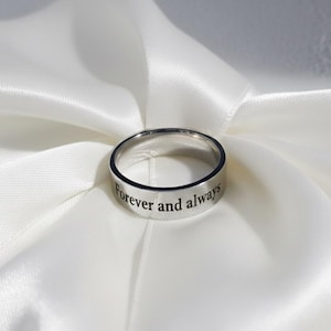 Peut inclure: Une bague argentée gravée des mots "Forever and always" sur la bande extérieure. La bague est posée sur un tissu de satin blanc, créant un contraste doux et élégant. La bague est de conception simple.
