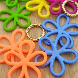 Könnte beinhalten: Eine Sammlung bunter blumenförmiger Anhänger in leuchtendem Gelb, Magenta, Limettengrün, Orange und Blau, begleitet von zwei kleinen goldfarbenen Creolen mit einer texturierten, gehämmerten Oberfläche.