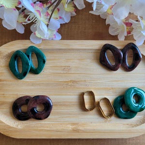 May include: A wooden tray displays several pairs of earrings. There are dark green, brown, and gold earrings. The earrings have various shapes, including geometric and hoop designs. White flowers are in the background.