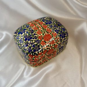 May include: A square decorative box with a floral pattern. The box has a cream base with red, blue, and green floral designs. The flowers are arranged in a repeating pattern across the top and sides of the box.