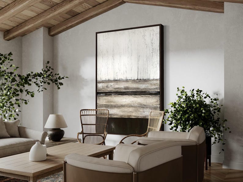 May include: A modern living room scene with a large abstract artwork in a dark frame. The room features neutral-toned furniture, including a sofa and chairs, a wooden coffee table, a lamp, and potted plants, creating a stylish and comfortable interior.