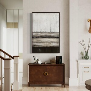 May include: An abstract landscape painting in a dark wood frame hangs above a dark brown cabinet with gold handles. The painting features textured white, grey, and black horizontal bands. The cabinet is adorned with decorative items, including candles and books.