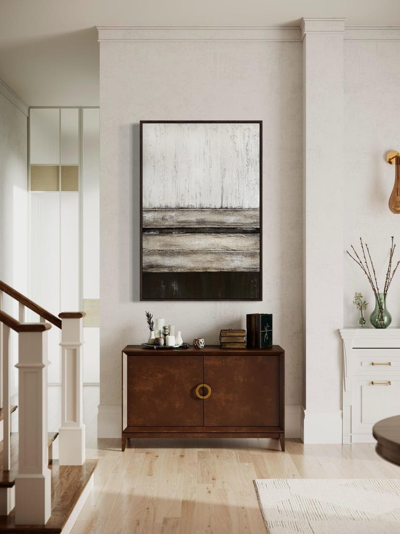 May include: An abstract landscape painting in a dark wood frame hangs above a dark brown cabinet with gold handles. The painting features textured white, grey, and black horizontal bands. The cabinet is adorned with decorative items, including candles and books.