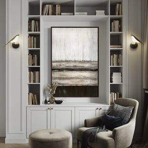 May include: A white built-in bookcase with an abstract painting, a grey armchair, and a round ottoman. The bookcase is filled with books and has two bird-shaped wall sconces. The painting has a white and brown colour scheme.