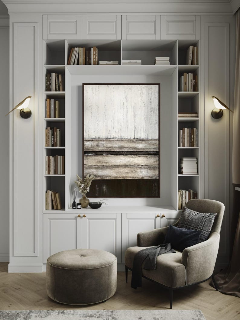 May include: A white built-in bookcase with an abstract painting, a grey armchair, and a round ottoman. The bookcase is filled with books and has two bird-shaped wall sconces. The painting has a white and brown colour scheme.