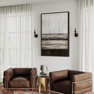 May include: Two brown leather armchairs with metal frames sit on a patterned rug. A small wooden side table with a bust sculpture is between the chairs. An abstract painting hangs on the wall, and sheer curtains frame the window.