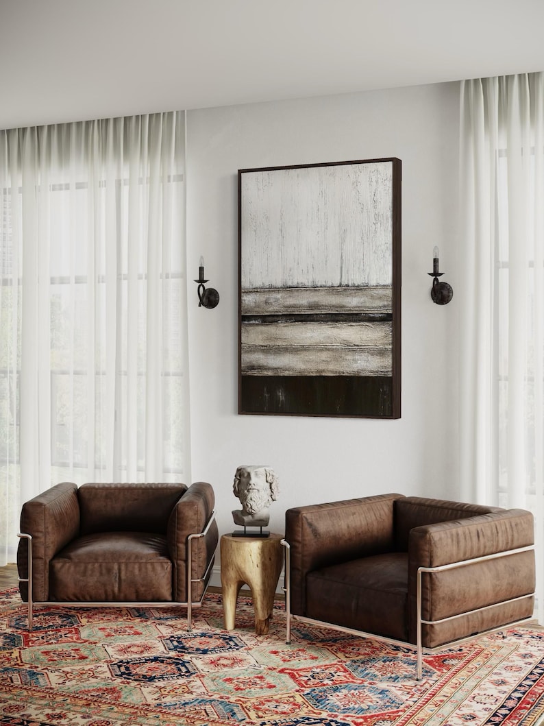 May include: Two brown leather armchairs with metal frames sit on a patterned rug. A small wooden side table with a bust sculpture is between the chairs. An abstract painting hangs on the wall, and sheer curtains frame the window.