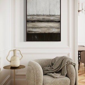 May include: Abstract framed wall art with a textured design in white, grey, and dark brown. A beige armchair with a grey knitted throw is in the foreground. A small side table with a vase is next to the chair. The room has wooden flooring.