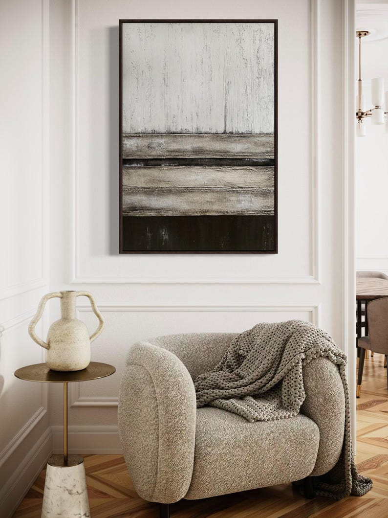 May include: Abstract framed wall art with a textured design in white, grey, and dark brown. A beige armchair with a grey knitted throw is in the foreground. A small side table with a vase is next to the chair. The room has wooden flooring.