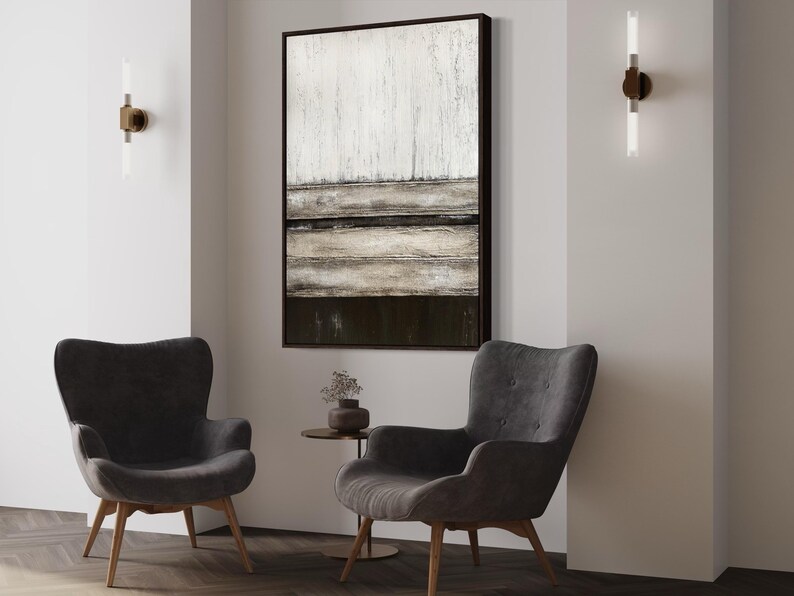 May include: A modern living room setup with two grey armchairs and a large abstract painting. The painting is framed in black and hangs above the chairs. Two wall lights with vertical tubes are mounted on the wall. A small side table is between the chairs.