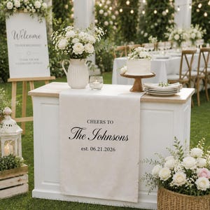 May include: A white wooden table draped with a beige runner featuring the black text CHEERS TO The Johnsons est. 06.21.2026. The table holds a white pitcher with white flowers, a small glass candle holder, a white cake on a stand, and a stack of plates.