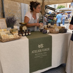 May include: A market stall table covered with a beige linen cloth featuring a dark green runner with the text ATELIER VERDE BOTANICAL SOAP HANDMADE IN PROVENCE. The table displays various rectangular soap bars in baskets and on display.