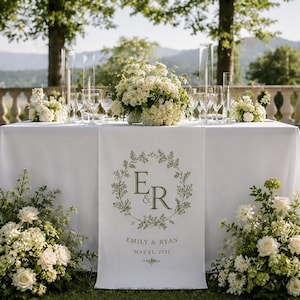 May include: A white fabric table runner featuring a centered olive green botanical wreath design enclosing the letters E and R, with the text Emily & Ryan and May 21, 2023 printed below, displayed on a table decorated with white floral arrangements.