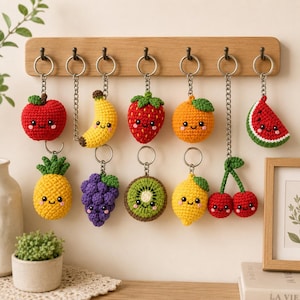May include: A collection of eleven crocheted fruit keychains hanging from a wooden rack. The fruits include an apple, banana, strawberry, orange, watermelon slice, pineapple, grapes, kiwi slice, lemon, and a pair of cherries, all with smiling faces.