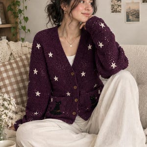 May include: A deep purple long-sleeve crochet cardigan featuring a V-neckline, four brown buttons, and two front pockets. The garment is decorated with white embroidered stars and a black cat motif on each pocket, paired with white wide-leg trousers.