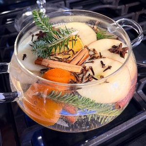Peut inclure: Une théière en verre transparent remplie d'eau, de tranches de pomme, d'orange, de bâtons de cannelle, de clous de girofle et de brins de conifères. La théière est posée sur une plaque de cuisson noire, suggérant une boisson chaude ou un pot-pourri.