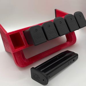 May include: A red 3D-printed magazine holder with five black magazine base plates and a black magazine. The holder has a unique design with a slot for a single magazine and angled slots for the base plates.