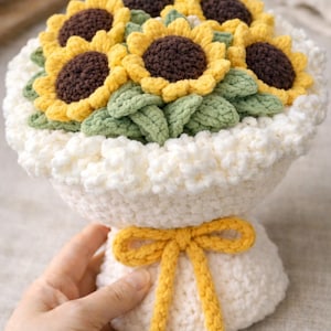 May include: A crocheted bouquet of sunflowers. The flowers have yellow petals and brown centers, with green leaves. The bouquet is in a white crocheted vase, tied with a yellow bow.
