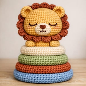 Peut inclure: Un jouet à empiler fait à la main au crochet représentant un lion avec un corps jaune, une crinière marron et les yeux fermés. Le lion est posé sur une pile de cinq anneaux colorés crème, vert, orange et bleu. Le jouet est pour enfants.