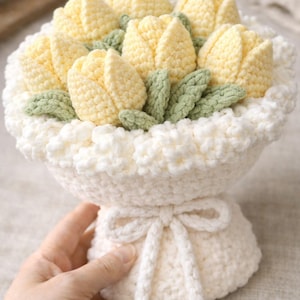 Op de afbeelding: Een handgemaakt gehaakt boeket met gele tulpvormige bloemen en groene bladeren, gerangschikt in een witte, gerimpelde mand. Het boeket is vastgebonden met een witte strik, met een zachte, getextureerde uitstraling.