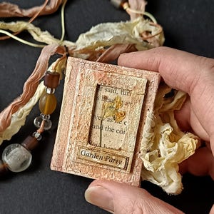 Puede incluir: Pequeña pieza de arte en capas, de color rosa, con las palabras "Garden Party" en una etiqueta rectangular. La pieza presenta una flor seca y texto de un libro. Está adornada con cuentas y cintas en tonos marrón y crema.