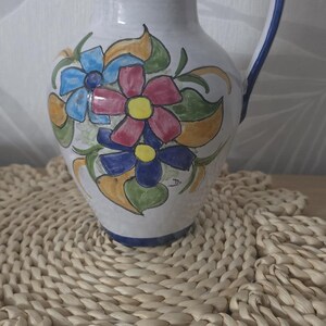 Op de afbeelding: Een witte keramische kan met een handgeschilderd bloemmotief in blauw, rood en geel, groene bladeren en een blauwe rand en handvat. De kan heeft een ronde buik, een smalle hals en een gebogen handvat dat aan de bovenrand is bevestigd.